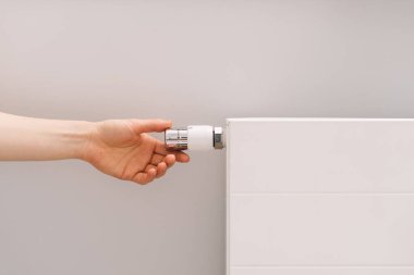 Tüketim ve enerji tasarrufu kavramı. Bir el, oturma odasındaki beyaz elektrikli ısıtıcının termostatını çalıştırarak modern bir apartman dairesinde soğuk mevsim günlerinde konfor sağlıyor..