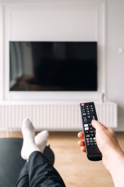 Vertical shot of living room features a large flat screen smart TV mounted on the wall. Person reclines with a remote controller, enjoying a comfortable and stylish interior perfect for entertainment.