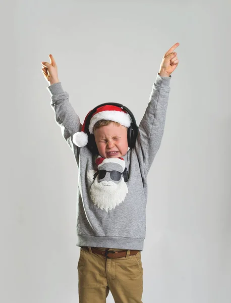 Happy kid in sweater of Canta Clause holding with raised hands. Christmas kid music star. Child waiting for present with closed eyes and listening music in headphones