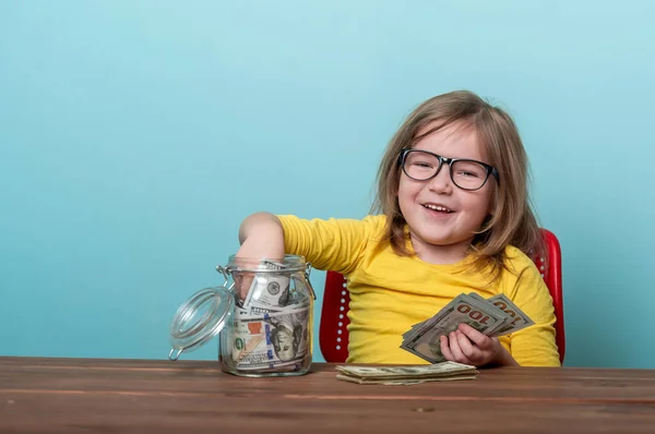 Happy child takes money from glass jar full of dollars. Portrait of kid with money in hand. Savings for future. Financial education for kids. How to think and investing and do not afraid