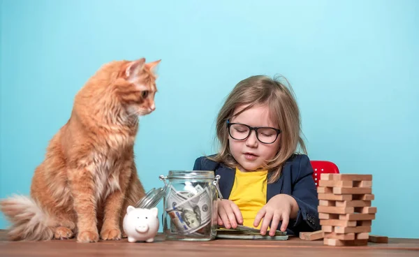 Смішний маленький бізнесмен і імбирний кіт рахують заощадження на столі. Освіта, як бути багатим. Дитина поклала гроші в банку і скарбничку — стокове фото