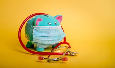 Mavi Piggybank 'in etrafındaki steteskop sarı arka planda izole edilmiş. Koruyucu sağlık maskeli domuz kumbarası.