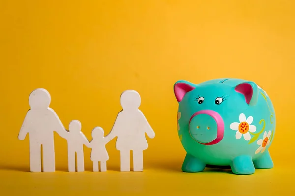 蓝色小猪银行，白色木制家庭图标。在黄色背景下孤立的家族人物和储蓄罐。储蓄和家庭财政预算。今后的节余. — 图库照片