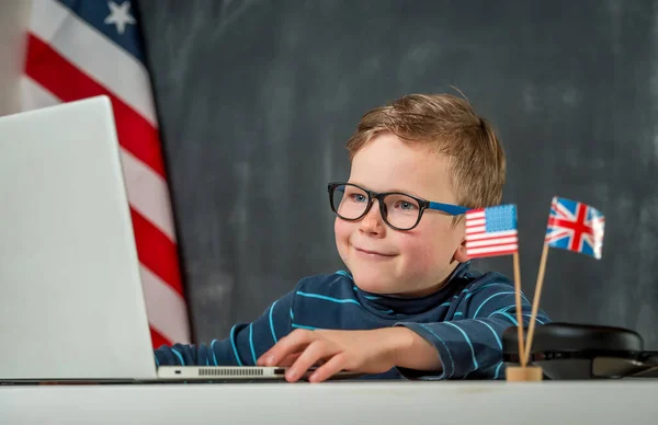 Возвращайся в школу. Милый и счастливый маленький мальчик имеет урок на ноутбуке. Английский язык на компьютере. Семилетний ребенок учится через систему электронного обучения онлайн. Флаг США и Великобритании — стоковое фото