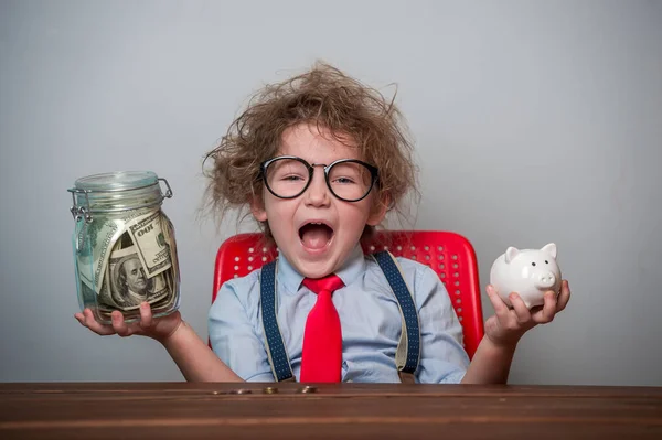 Kind en geld. Grappig opgewonden kind speelt met geld. Opleiding voor kleine zakenman hoe rijk te worden als je groot bent. Leerling in bril en stropdas met spaarvarken en pot vol geld — Stockfoto