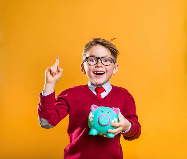 Menino da escola feliz com dinheiro apontando para cima isolado em amarelo. Homem de negócios elegante fresco. Como ser rico. Retrato de um menino nerd bonito carregando um banco porquinho com suas economias. Sucesso, motivação, vencedor. — Fotografia de Stock