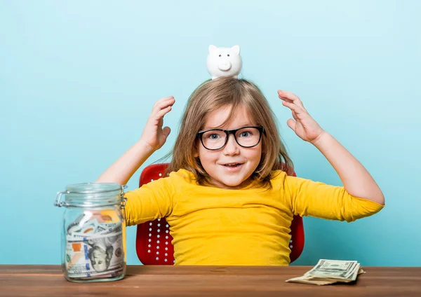 Cute little girl in eyeglasses and bright yellow shirt shaking piggy box on blue background. Happy kid and money — Stock Photo, Image