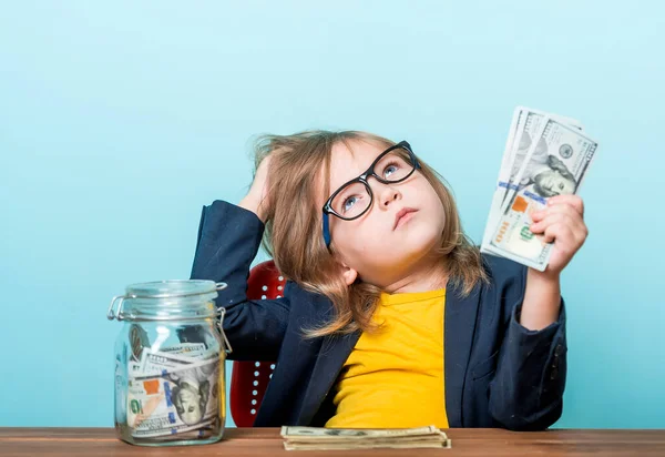 Marzycielka przy stole liczy pieniądze na niebieską ścianę. Dzieciak w okularach marzy o przyszłości. Edukacja do liczenia. Darowizna na cele charytatywne — Zdjęcie stockowe