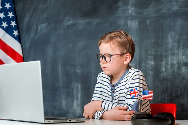 Онлайн урок английского языка. Красавчик учит уроки на ноутбуке. Умный ботаник в очках. Ребенок в наушниках изучает английский онлайн. — стоковое фото