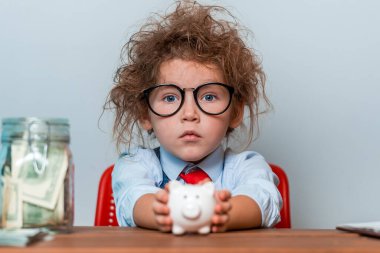 Akıllı çocuk kumbaraya para koyuyor. Domuz kumbaralı gözlüklü kız çocuğu. Para ve eğitim