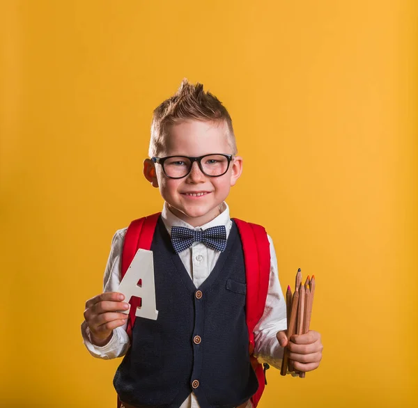Портрет улыбающегося школьника с рюкзаком, держащего карандаши на желтом фоне. Мальчик в очках с красной сумкой. Возвращение в школу. — стоковое фото