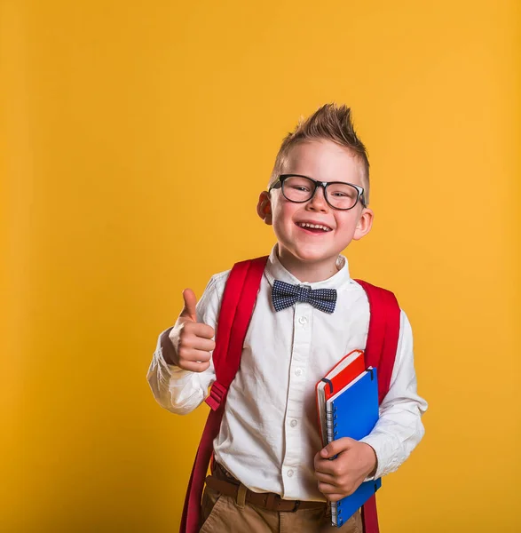 Porträtt av glad leende pojke på färg bakgrund. Snygg blond pojke i glasögon med röd ryggsäck och anteckningsbok och böcker. — Stockfoto