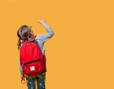 Back view of pupil in red backpack pointing up with the finger. childhood, education and people concept. happy smiling student girl with school bag go to school. Stylish kid. Child isolated on yellow.