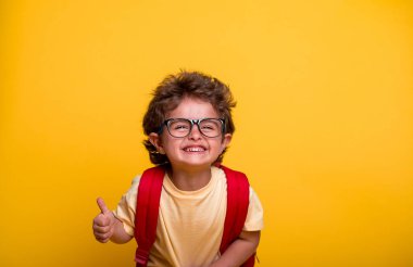 Portrait of happy laughing little kid boy isolated on yellow background. Back to school. Preschool boy in kindergarten. Smartchild in glasses with red backpack. Educate at home or daycare