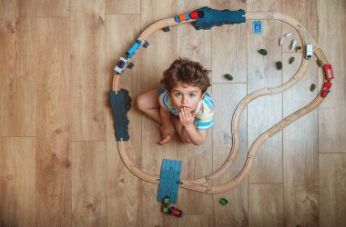 Oyuncak tren yolu oynayan çocuğun en iyi görüntüsü. Çocuk tahta oyuncaklarla yerde oturuyor. Küçük çocuk için oyuncaklar. Anaokulu demiryolu inşaatı ve evdeki bloklar, kreş. Anaokulu eğitim oyunları