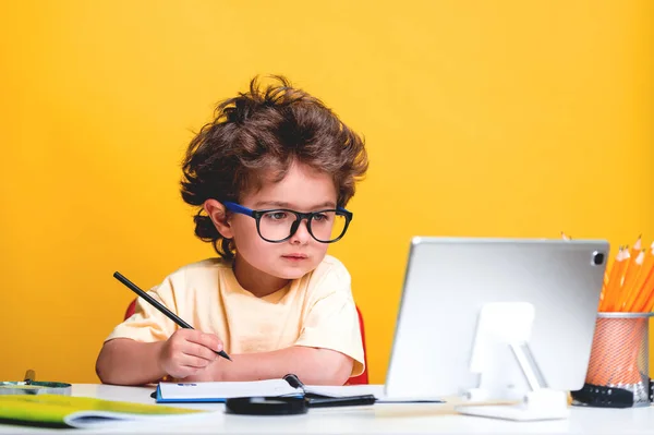 Niño inteligente sentado en su escritorio con computadora portátil. Alumno con lápiz en dibujo a mano aislado sobre fondo amarillo. Divertido niño pequeño haciendo tareas de escritura y lectura en casa —  Fotos de Stock