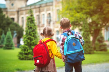 Sırt çantalı iki küçük öğrenci el ele okula gidiyor. Okul bahçesinde birinci sınıflar. Çocuklar okula geri döner. Kız ve erkek öğrenciler ilkokula gider..