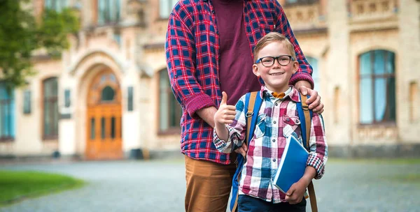 Το χαρούμενο χαμογελαστό αγόρι με τα γυαλιά με τον αντίχειρα σηκωμένο θα πάει σχολείο για πρώτη φορά. Παιδί με σχολική τσάντα και βιβλίο στο δρόμο για το κτίριο. Σίγουρο παιδί. Ο μπαμπάς υποστηρίζει τους μαθητές. Πίσω στο σχολείο.. — Φωτογραφία Αρχείου