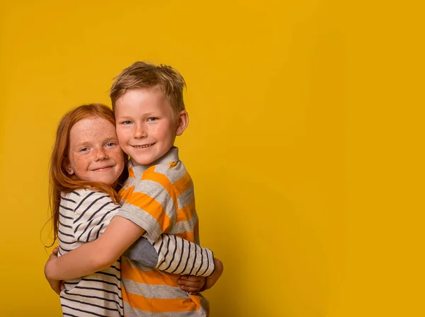 Happy ginger kids girl and boy hugging isolated on yellow background. Freckled red hair child in striped shirt.brother and sister smiling and embracing. Summer camp for children.