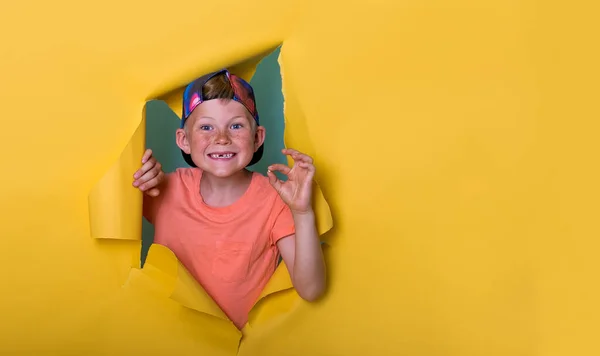 Funny smiling seven years old kid boy breaks through yellow paper wall. Happy school child holding missing tooth. Back to school — Stock Photo, Image