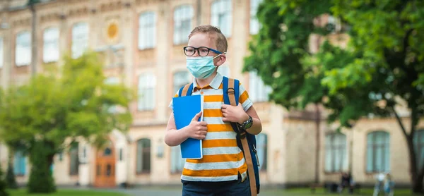 Schoolboy in face mask and with backpack go back to school. Corona virus outbreak. Child in glasses with notebook after covid-19 quarantine. Kid boy in safety mask for virus prevention. Smart pupil