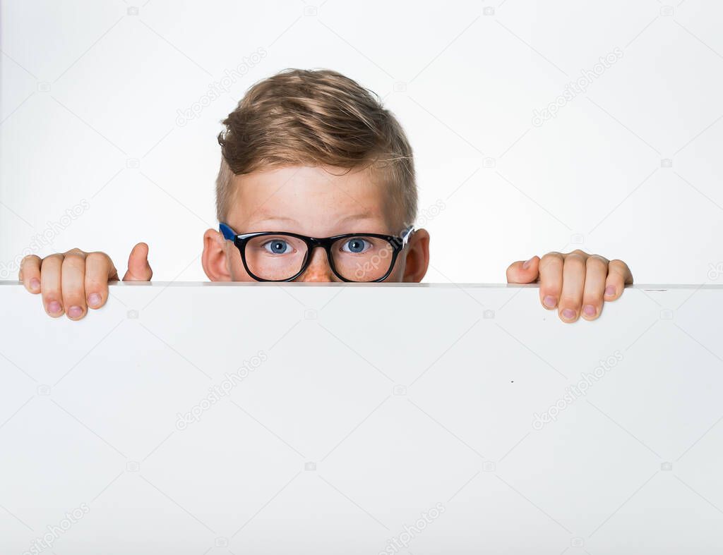 Portrait of surprised child boy in glasses hiding blank white poster or placard. Advertisement or sale concept. School kid peeking out board free space for logo, design or message. Hiding face