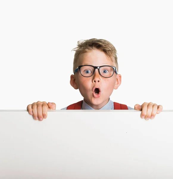 Kleine schooljongen met bril verschijnt achter spandoek. Verbazingwekkend kind dat een witte poster of plakkaat verstopt. Reclame of verkoop. De leerling gaat terug naar school in uniform. kind boven leeg reclamebord — Stockfoto