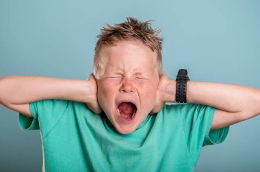 Küçük okul çocuğu kulaklarını elleriyle kapatıyor, mavi arka planda izole edilmiş. Çocuk gözleri kapalı ve ağzı açık bağırıyor. Kulak ağrısı, diş ağrısı. Ache. Stres yüzünden. Dinlemek ve duymak istemiyorum.