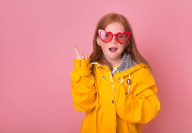 Kırmızı kalp gözlüklü neşeli kız çocuğu hava tahmininde parmağıyla işaret ediyor. Sarı su geçirmez yağmurluk ve lastik çizmeli çilli kızıl saçlı kız. Her türlü havada aşk.