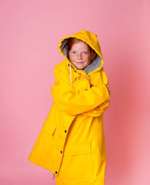 Sarı yağmurlukla kaplı neşeli, komik liseli kız. Yağmurlu sonbahar günlerinde korunma. Pembe arka planda izole edilmiş büyük giysili şık bir akraba.