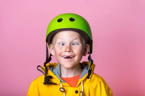 Funny kid boy portrait. crazy comic blond child makes grimace and crosses eyes. school boy with broken front tooth playing fool in green protect helmet and yellow coat. bright people face expressions. — Stock Photo, Image