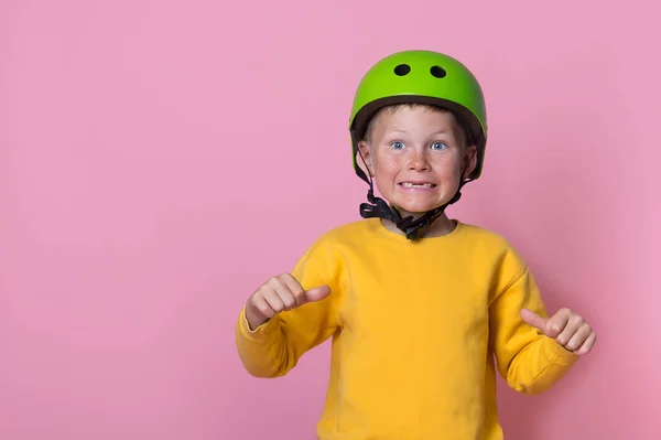 Charming blond kid boy in bright green helmet and yellow clothes with thumbs up pointing on him. School kid isolated on pink. Funny face. Portrait of stylish 7 years old kid. Space for text on shirt — Stock Photo, Image