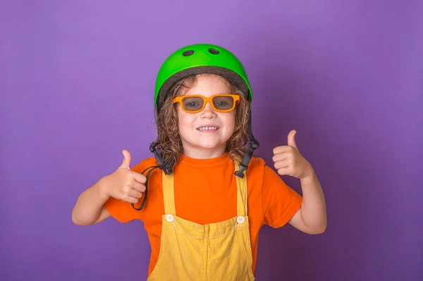 Funny kid portrait in green helmet and orange glasses gives thumbs up. blonde european girl with smiling faces isolated on violet background. Safety and protection concept. — Stock Photo, Image