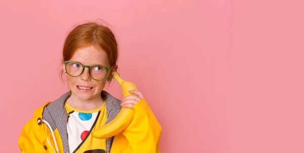 一个戴着绿色眼镜、大眼睛惊讶的戴眼镜的女学生.穿着黄色夏衣的红头发小女孩在香蕉上聊天，就像在电话里一样，背景是粉色的，有复制的空间 — 图库照片