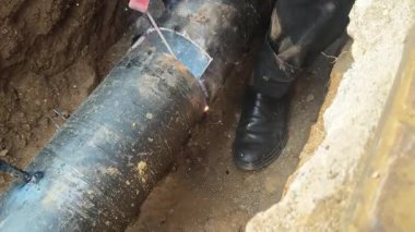 Bir adam kalın bir demir boruyu kaynaklıyor. İşçi, kısmen toprağa gömülü büyük, koyu renkli bir boruya bir yama kaynak yapıyor. Siperde çalışırken kıvılcımlar ve duman görünür..