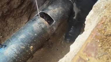 Bir adam kalın bir demir boruyu kaynaklıyor. İşçi, kısmen toprağa gömülü büyük, koyu renkli bir boruya bir yama kaynak yapıyor. Siperde çalışırken kıvılcımlar ve duman görünür..