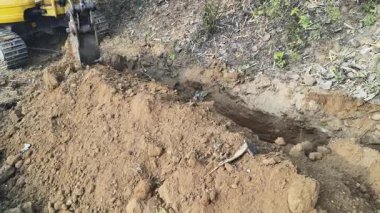 Bir kazıcı, boru döşemek için dar bir çukur kazmak için kovasını kullanır. Çalıların ve bitkilerin yanındaki kuru, kahverengi toprağı kazıyor. Su boru hattı döşeme çalışmaları devam ediyor..