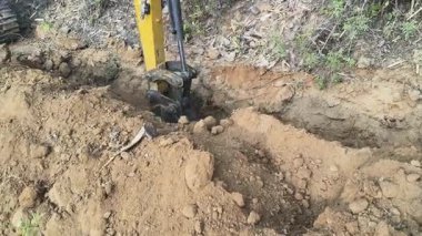 Bir kazıcı, boru döşemek için dar bir çukur kazmak için kovasını kullanır. Çalıların ve bitkilerin yanındaki kuru, kahverengi toprağı kazıyor. Su boru hattı döşeme çalışmaları devam ediyor..