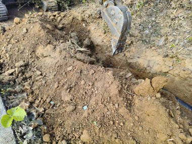 Bir kazıcı, boru döşemek için dar bir çukur kazmak için kovasını kullanır. Çalıların ve bitkilerin yanındaki kuru, kahverengi toprağı kazıyor. Su boru hattı döşeme çalışmaları devam ediyor..