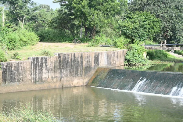 Küçük bir beton baraj nehri kaplar. Su merkezden akar ve beyaz bir köpük oluşturur. Ön ve yan tarafları çimen ve yeşil yapraklarla sınırlıdır. Sahne huzur dolu..