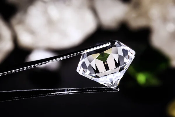 cut diamond held by a jeweler\'s tweezers, with rough diamond stones in the background, spot focus