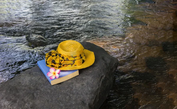 黄色的妇女帽子和书在瀑布的石头上 — 图库照片