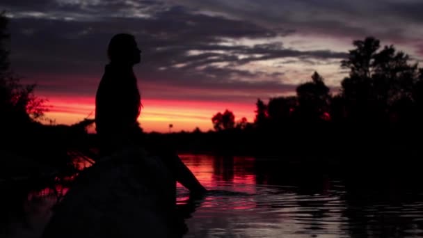 Дівчина сидить біля річки на заході сонця — стокове відео