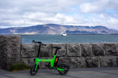 REYKJAVIK, ICELAND - 11 Haziran 2021: Reykjavik limanında yeşil elektrikli bisiklet. Arka plandaki körfezde demirlemiş lüks bir yat ve Esja Dağı sırtı manzarası.. 