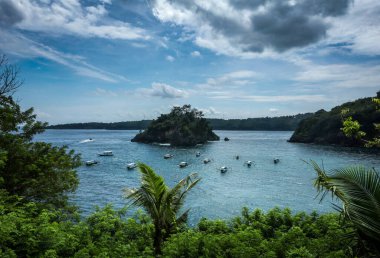 Kuzeybatı Nusa Penida, Bali, Endonezya 'daki Pandan Sahili. Ortadaki küçük ada, körfeze demirlemiş tekneler tropikal yağmur ormanlarıyla çevrili.. 