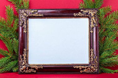 Mockup. Christmas. Ornate wooden picture frame with intricate detailing placed among lush evergreen branches, creating festive atmosphere against bright red background, perfect for seasonal displays