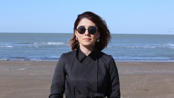 Depressed young woman near the sea in a windy day. Beautiful young brunette woman outdoors portrait near the sea. Young pretty girl in black dress walking near the seaside at sunset. — Stock Video