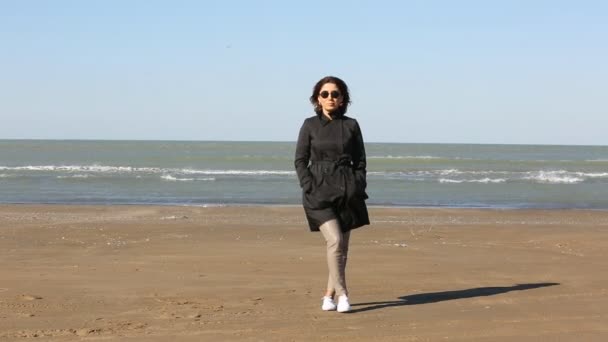 Mujer joven deprimida cerca del mar en un día ventoso. Hermosa mujer morena joven al aire libre retrato cerca del mar. Joven chica bonita en vestido negro caminando cerca de la playa al atardecer . — Vídeos de Stock