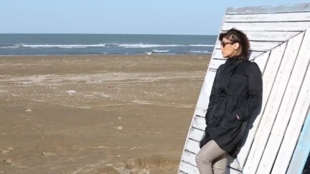 Deprimerade unga kvinna nära havet i en blåsig dag. Vacker ung brunett kvinna utomhus porträtt nära havet. Ung söt flicka i svart klänning promenader vid havet i solnedgången. — Stockvideo