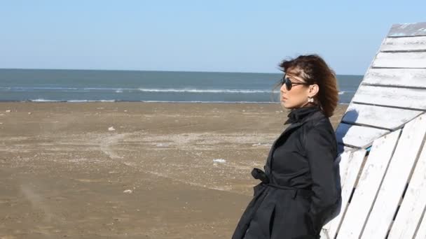 Depressed young woman near the sea in a windy day. Beautiful young brunette woman outdoors portrait near the sea. Young pretty girl in black dress walking near the seaside at sunset. — Stock Video